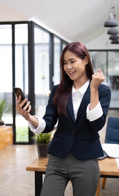 Fröhliche, erstaunte erfolgreiche junge asiatische Geschäftsfrau, die mit einem Smartphone im Büro steht und eine überraschte Nachricht erhält