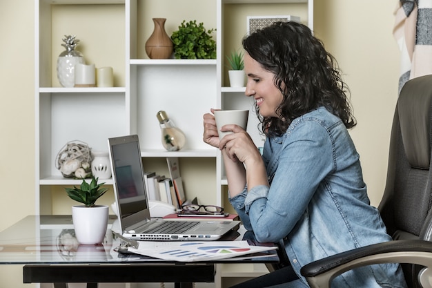 Fröhliche erwachsene Frau, die aromatischen Kaffee trinkt und Laptop zu Hause benutzt