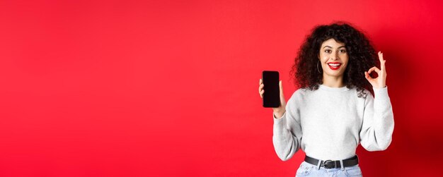 Foto fröhliche europäische frau mit lockigem haar, die einen leeren handy-bildschirm und eine okay-geste zeigt, die s lächelt