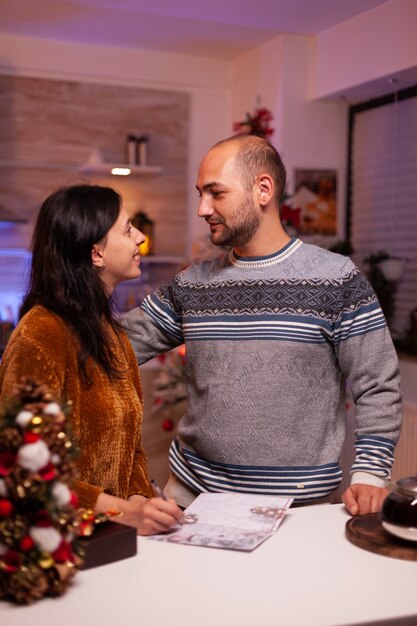 Fröhliche Familie, die Weihnachtsgrußpostkarte in der weihnachtlich dekorierten Küche schreibt