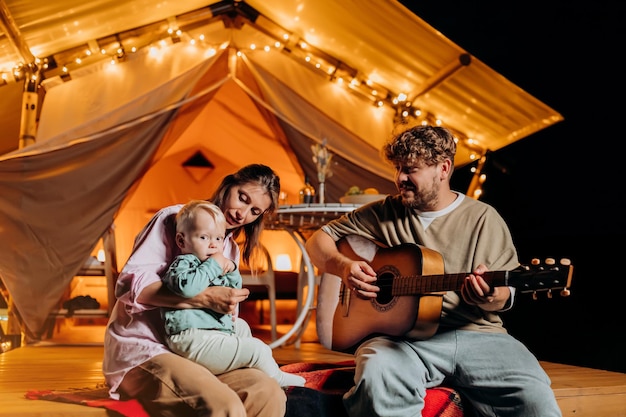 Fröhliche Familie entspannt sich und verbringt Zeit zusammen im Glamping am Sommerabend und spielt Gitarre in der Nähe des gemütlichen Lagerfeuers Luxus-Campingzelt für Erholung und Erholung im Freien Lifestyle-Konzept