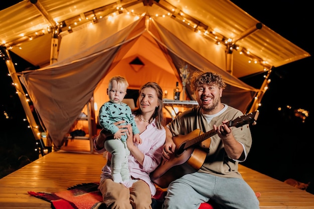 Fröhliche Familie entspannt sich und verbringt Zeit zusammen im Glamping am Sommerabend und spielt Gitarre in der Nähe des gemütlichen Lagerfeuers Luxus-Campingzelt für Erholung und Erholung im Freien Lifestyle-Konzept
