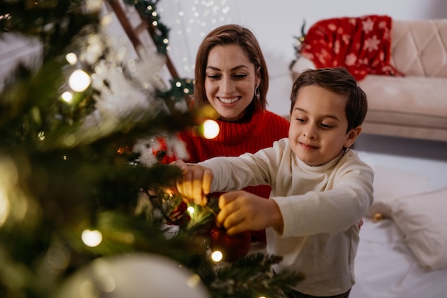 Fröhliche festliche Familie von Frauen mit kleinem Sohn, der den Weihnachtsbaum mit Lichtern und Spielzeug schmückt