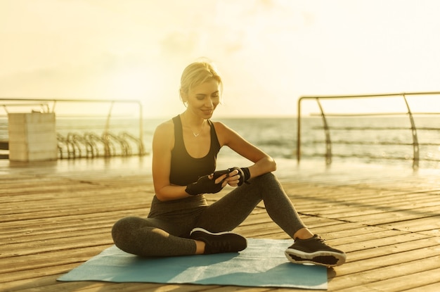 Fröhliche Fitnessfrau sitzt auf Matte und benutzt ein Smartphone am Strand
