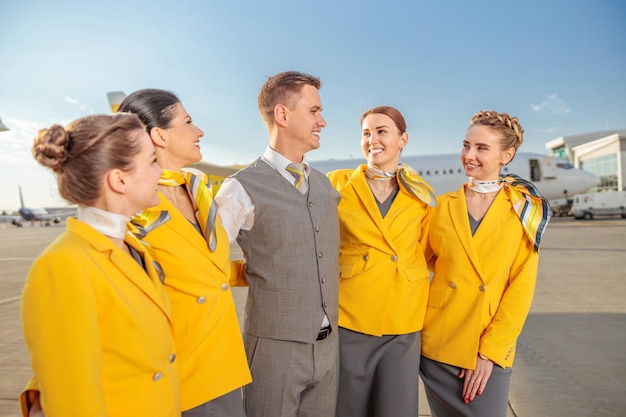 Fröhliche Flugbegleiterinnen schauen männliche Kollegen an und lächeln, während sie unter blauem Himmel am Flughafen stehen
