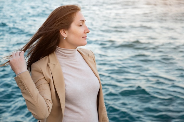 Fröhliche Frau berührt langes Haar, das am Wasser steht und die schöne Natur gegen das plätschernde Meer genießt