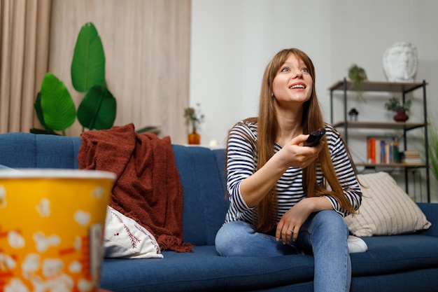 Fröhliche Frau, die auf dem Sofa sitzt, wechselt die Kanäle auf dem Fernseher