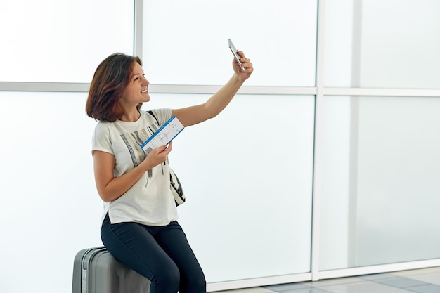 Fröhliche Frau, die selfie mit Ticket im Flughafen vor Abflug auf weißem Fenster nimmt