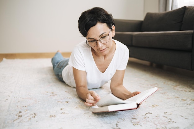 Fröhliche Frau, die zu Hause Papierbuch liest Brünette mit kurzen Haaren in Freizeitkleidung