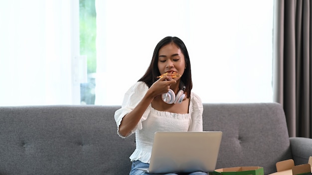 Fröhliche Frau im Kopfhörer, der Laptop verwendet und Pizza isst, während auf Sofa im Wohnzimmer sitzend.