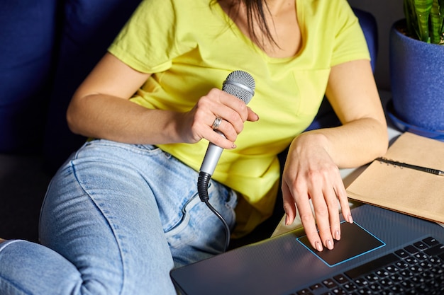 Fröhliche Frau in Freizeitkleidung Aufnahme eines Podcasts, der in ein Mikrofon mit Kopfhörern und Laptop, Notizbuch spricht
