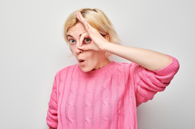 Fröhliche Frau in rosa Pullover macht ein OK-Zeichen mit der Hand über dem Auge und lächelt auf einem hellen Hintergrund