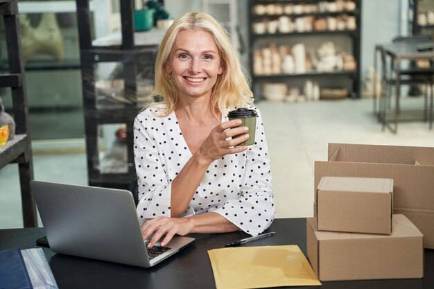 Foto fröhliche frau, kleinunternehmerin, die in die kamera lächelt, kaffee trinkt, während sie am laptop arbeitet