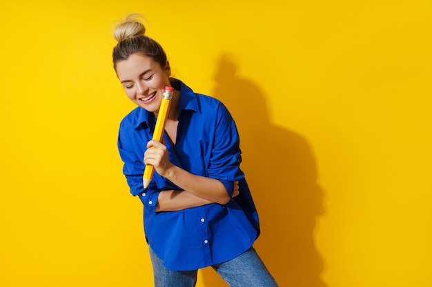 Fröhliche Frau mit blauem Hemd und riesigem Bleistift auf gelbem Hintergrund