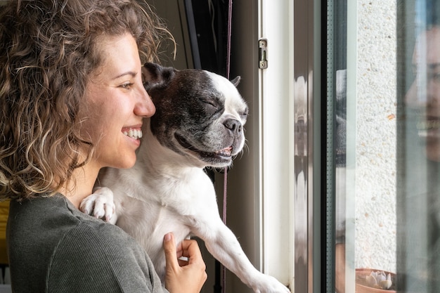 Fröhliche Frau mit Bulldogge, die zu Hause lächelt. Horizontale Ansicht der Frau mit Babyhund im Fenster