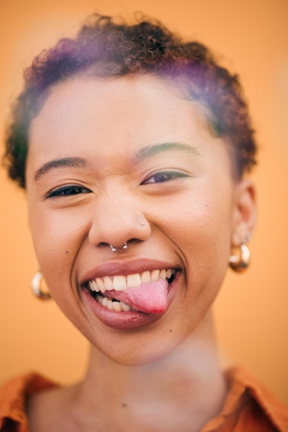 Fröhliche Frau mit herausgestreckter Zunge im Studio mit Lächeln und Selbstvertrauen, die sich albern fühlt. Orange Hintergrund, junges Porträt und afrikanische Frau mit trendiger moderner und studentischer Mode mit Gen-Z-Glanz
