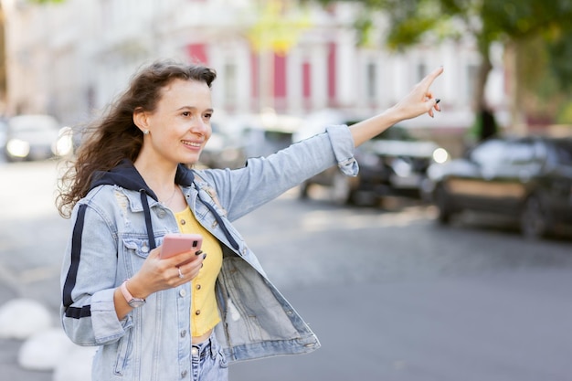 Fröhliche Frau mit lockigem Haar in Jeansjacke fängt an einem hellen sonnigen Tag ein Auto in der Stadt
