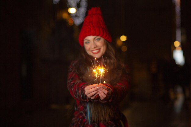 Fröhliche Frau mit rotem Hut, die sich abends mit funkelnden Lichtern im Freien amüsiert