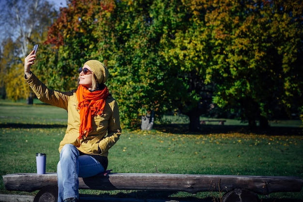 Fröhliche Frau mit Sonnenbrille, die auf einer Holzbank im Herbstpark sitzt und ein Selfie auf dem Smartphone macht. Verschwommener grüner Hintergrund, Kopierbereich.