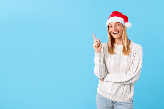 Fröhliche Frau mit Weihnachtsmannshut zeigt auf den freien Raum auf blauem Hintergrund