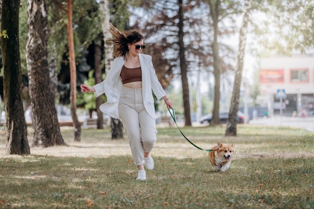 Fröhliche Frau mit weißem Anzug und Sonnenbrille, die mit dem walisischen Corgi Pembroke-Hund in der Parklandschaft der Stadt spazieren geht