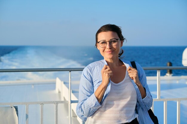 Fröhliche Frau mittleren Alters auf dem Deck des Kreuzfahrtschiffes, Kopienraum, blauer Himmel, Sonnenuntergang am Meer. Lächelnde reife Frau, welche die Kamera betrachtet