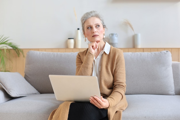 Fröhliche Frau mittleren Alters mit Laptop beim Sitzen auf dem Sofa zu Hause.