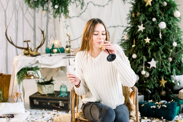 Fröhliche Frau sitzt in der Nähe des Weihnachtsbaumes und hält Bengal Licht und trinkt Champagner