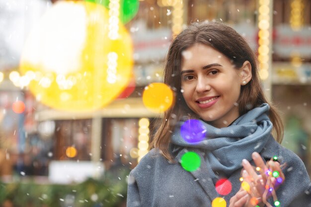 Fröhliche Frau trägt einen grauen Mantel, der während des Schneefalls mit Bokeh-Licht auf die Straße geht. Freiraum