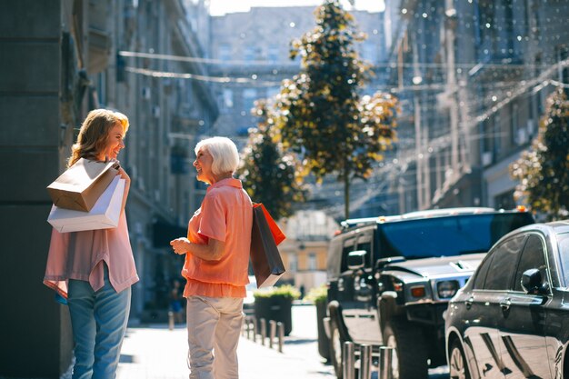 Fröhliche Frau und ihre Mutter, die Papiertüten tragen und sich im Freien anlächeln