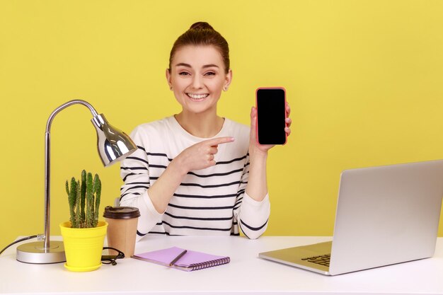 Fröhliche Frau zeigt auf Smartphone mit leerem weißen Display und prahlt mit neuem Modell