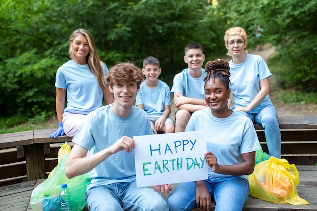 Fröhliche Freiwillige, die ein Plakat mit der Botschaft „Happy Earth Day“ halten. Freiwilligenarbeit für wohltätige Zwecke, die Menschen und das Ökologiekonzept reinigen. Gruppe glücklicher Freiwilliger mit Müllsäcken, die den Bereich im Park reinigen