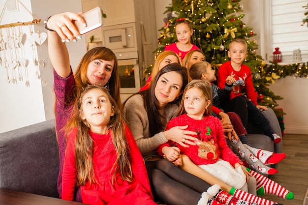 Fröhliche Freunde, die sich auf dem Smartphone selbst machen und lächeln. Sie sitzen vor dem Weihnachtsbaum und umarmen ihre Kinder