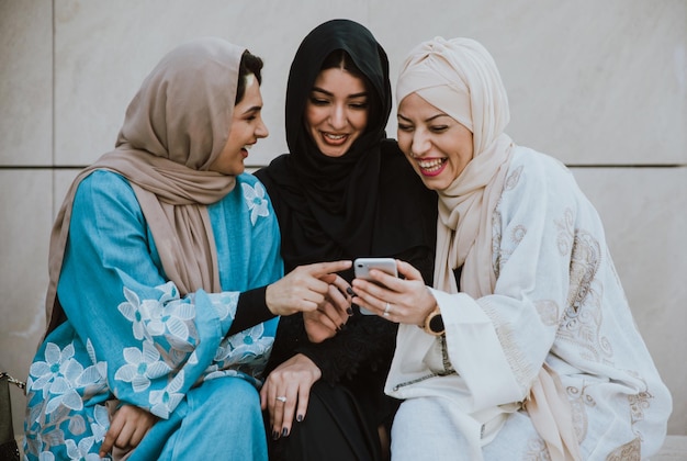 Fröhliche Freunde im Hijab sitzen an der Wand und schauen sich das Telefon an.