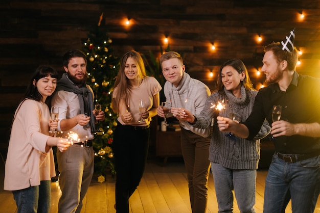 Fröhliche Freunde posieren mit Champagnergläsern und mit bengalischen Lichtern und feiern Neujahr. Weihnachtsbaum mit Girlande im Hintergrund. Freunde feiern Heiligabend.