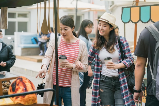 Fröhliche Freundinnen teilen sich an Wochenenden Zeit im Freien auf dem Kreativmarkt. junge freundinnen, die kaffee zum mitnehmen halten und in der sommersaison auf den verkäufer zeigen. fröhliche frauen, die spaß haben.