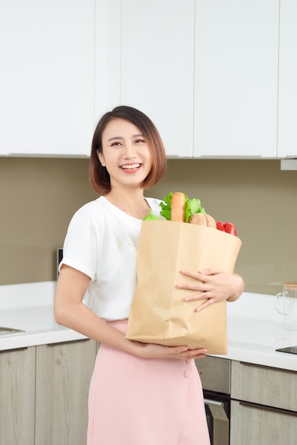 Fröhliche, fröhliche Asiatin, die eine Papiertüte voller frischer Produkte, Gemüse, Grüns, Baguette und Bananen hält.