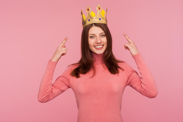 Fröhliche, fröhliche brünette Frau in rosafarbenem Pullover, die mit den Fingern auf die goldene Krone auf ihrem Kopf zeigt und ihr Führungsqualitäten-Konzept der Autorität zeigt Studioaufnahme isoliert auf rosafarbenem Hintergrund