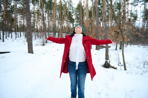 Fröhliche, fröhliche Frau, die die Arme hochhebt, während sie in einer schneebedeckten Natur wirbelt und einen schönen kalten Wintertag genießt, während sie auf einem verschneiten Kiefernwald spazieren geht Wunderbares kühles Winterwetter