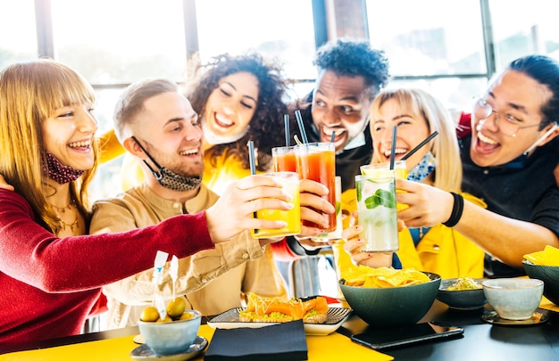 Fröhliche gemischtrassige Freunde, die Gesichtsmasken tragen und Cocktails im Bar-Restaurant anstoßen