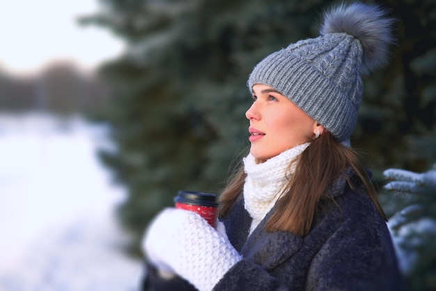 Fröhliche, gemütliche Frau, die draußen im Wald am verschneiten Wintertag heißen Tee oder Kaffee trinkt