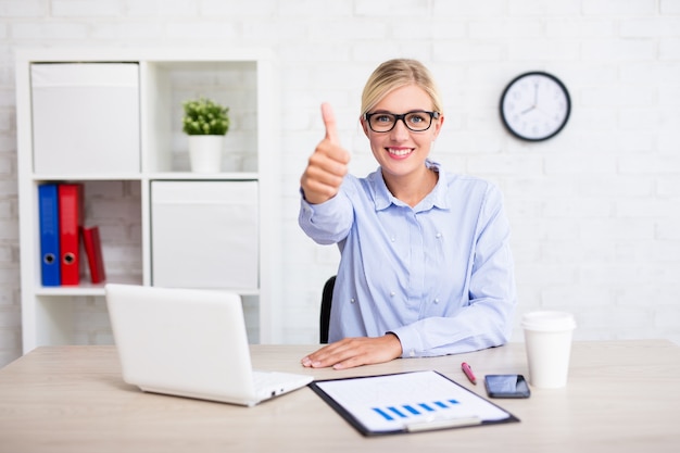 Fröhliche Geschäftsfrau, die im modernen Büro sitzt und Daumen hoch