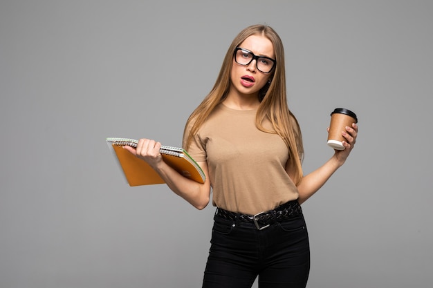 Fröhliche glückliche Frau mit zahnigem Lächeln, trägt Kaffee zum Mitnehmen und rotes Buch, freut sich, das Studium isoliert über der Studiooberfläche zu beenden Menschen-, Freizeit- und Trinkkonzept