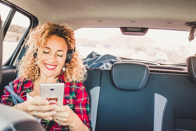 Fröhliche glückliche Frau setzt sich auf den Rücksitzen in einem modernen Auto und benutzt ein Mobiltelefon, um Musik mit Kopfhörern zu hören