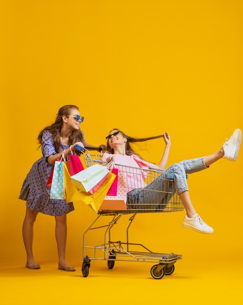 Fröhliche, glückliche Mädchen mit vielen Einkaufstüten, die auf einem Einkaufswagen sitzen, isoliert über gelbem Studiohintergrund