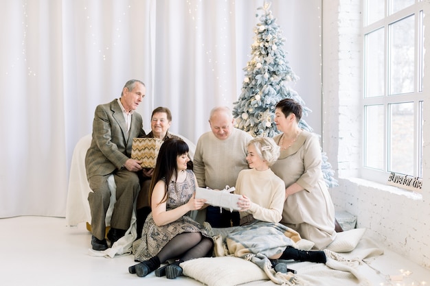 Fröhliche große Familie, die Weihnachtsgeschenke öffnet, während sie im gemütlichen Raum mit Tannenbaum sitzt