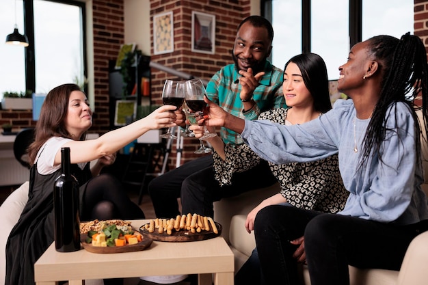 Fröhliche Gruppe multiethnischer Menschen, die auf einer Weinparty anstoßen, während sie ein Freundschaftsereignis feiern. Fröhliche, vielfältige Freunde, die auf der Geburtstagsfeier im Wohnzimmer mit Weingläsern klirren.