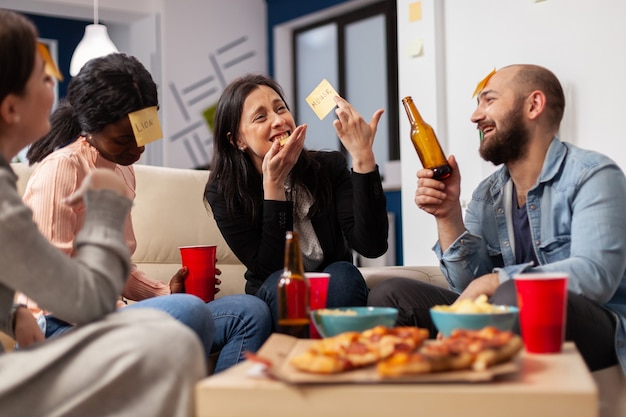 Fröhliche Gruppe von Kollegen, die nach der Arbeit im Büro ein Spiel von Scharaden genießen. Multiethnische Menschen spielen ein Nachahmungskonzept für unterhaltsame Aktivitäten, während sie Pizza essen und Bier trinken