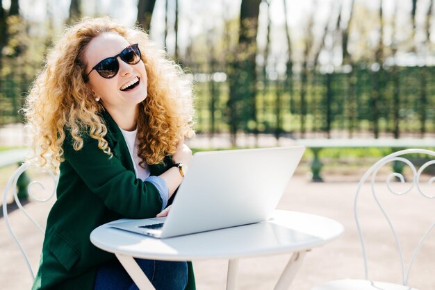 Fröhliche, hübsche blonde Frau mit lockigem Haar, Sonnenbrille und grüner Jacke, die sich im Park ausruht, am weißen Schreibtisch sitzt und mit ihrem Laptop online mit Freunden kommuniziert. Menschen-Emotionen-Konzept