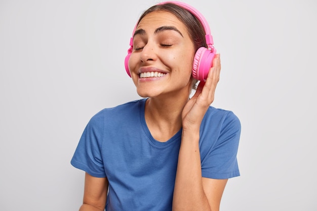 Fröhliche hübsche Frau mit dunklem Haar hält die Augen geschlossen und lächelt positiv genießt den Lieblings-Audiotrack in drahtlosen rosa Kopfhörern trägt blaues T-Shirt hört Lied isoliert über weißer Wand